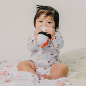 Baby sitting on a white blanket with a soft toy, wearing a patterned onesie.