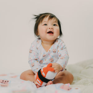 Baby sitting on a white blanket holding a plush toy sushi with a plain background